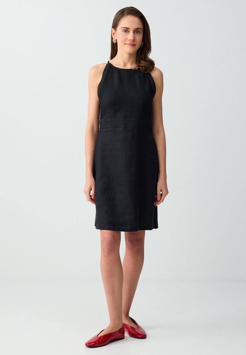 Femme debout portant une robe noire sans manches jusqu'au genou et des ballerines rouges sur un fond gris clair uni.