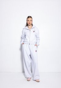 Graue Kapuzenjacke mit einem rosa Logo, kombiniert mit grauen, lockeren Sweatpants. Glatte Textur, lässiges Design und vordere Taschen.