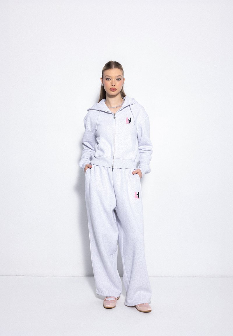 Graue Kapuzenjacke mit einem rosa Logo, kombiniert mit grauen, lockeren Sweatpants. Glatte Textur, lässiges Design und vordere Taschen.
