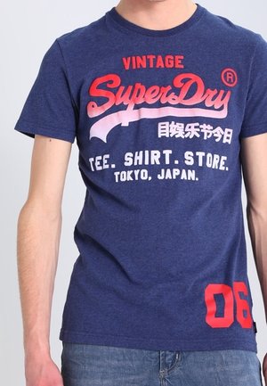 T-shirt bleu marine avec texte rouge et blanc "Vintage SuperDry", caractères japonais, et impression "Tee. Shirt. Store. Tokyo, Japon.", porté par une personne.