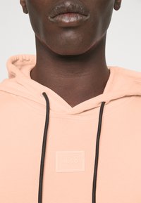 Kapotová mikina z jemné broskvové látky s logem, nastavitelné černé stahovací šňůrky a žebrovanými detaily kolem výstřihu.