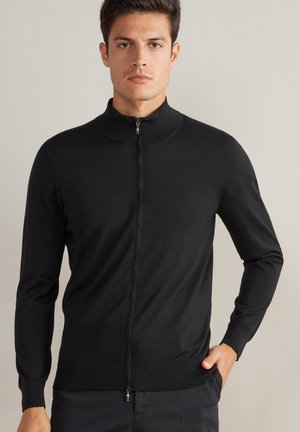 Jeune homme aux cheveux bruns courts portant un pull noir à fermeture éclair avec col montant, debout, une main dans la poche, sur un fond neutre.