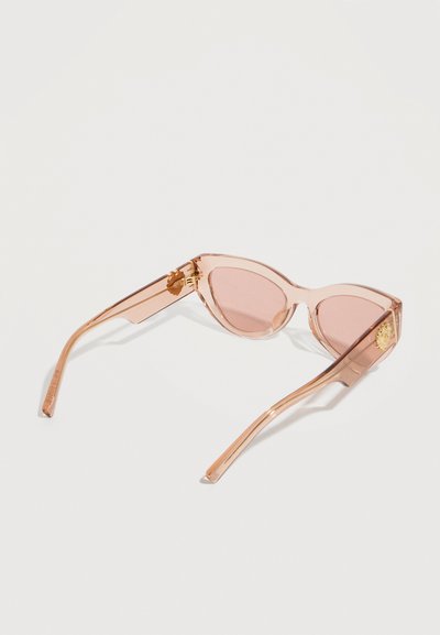 Ροζ διαφανή γυαλιά ηλίου σε σχήμα γατίσιου ματιού με χρυσό έμβλημα στα πλαϊνά, τοποθετημένα με την πρόσοψη προς τα κάτω σε λευκή επιφάνεια.