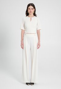 Jumpsuit bianco con top aderente, dettaglio a laccio e pantaloni a gamba larga. Tessuto liscio con delicato lucido, abbinato a scarpe nere a punta.