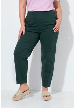 Frau trägt dunkelgrüne High-Waist-Hose mit seitlichen Knöpfen und beige Slip-On-Plateausandalen, steht vor weißem Hintergrund.