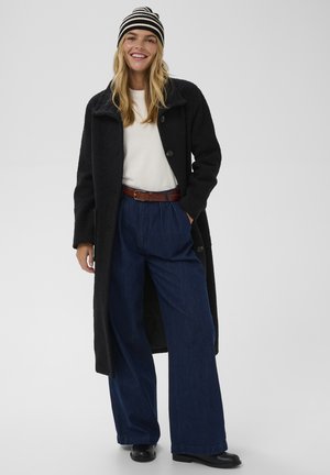 Long black coat with textured fabric, two large buttons, paired with wide blue denim trousers, a white top, and a striped beanie.