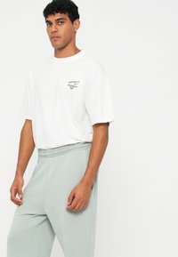 Weißes Baumwoll-T-Shirt mit kurzen Ärmeln und einem kleinen schwarzen Logo. Kombiniert mit hellgrünen, lässig geschnittenen Jogginghosen mit einem elastischen Bund.