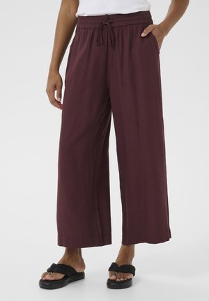 Kobieta ubrana w luźne bordowe szerokie spodnie z elastycznym pasem i sznurkiem, w połączeniu z czarnymi klapkami na płaskim obcasie z kokardką.