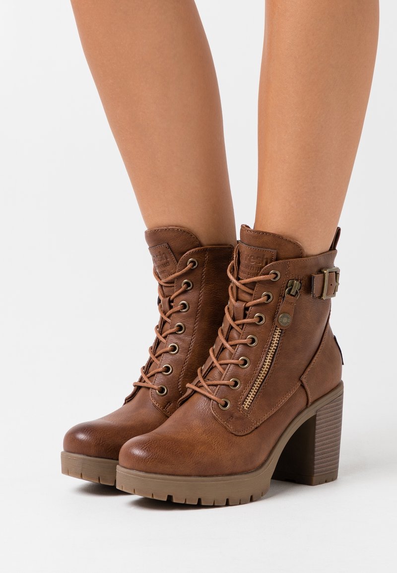 Bottes marron à lacets montantes avec fermeture éclair latérale et détail boucle, portées aux pieds avec talons carrés et semelles robustes.