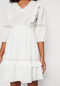 Vestido blanco con ribete de encaje, detalles con volantes en las mangas y la falda, mangas abullonadas tres cuartos y cintura fruncida en una persona.