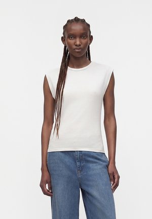 Vrouw met lange vlechten die een mouwloos wit topje en blauwe jeans met wijde pijpen draagt, staand tegen een effen witte achtergrond.