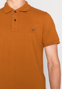 Polo w kolorze rdzy wykonany z bawełny, z teksturowanym materiałem, trzema guzikami przy dekolcie oraz małą łatą z logo na lewej piersi.