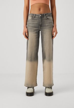 Persoon die tweekleurige vervaagde grijze en beige wijde jeans draagt, witte laarzen en een cropped perzikkleurige top, staand tegen een effen achtergrond.