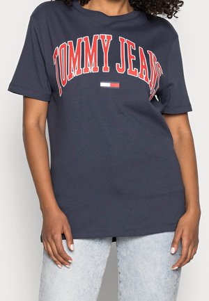 Personne portant un t-shirt bleu marine avec le texte "TOMMY JEANS" en rouge et blanc ainsi qu'un petit logo Tommy Hilfiger, associé à un jean délavé clair.
