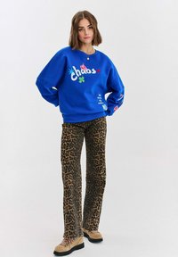 Modrá mikina s textom "chaos" a farebnými akcentmi, skombinovaná s hnedými širokými nohavicami s leopardím vzorom a béžovými šnurovacími topánkami.