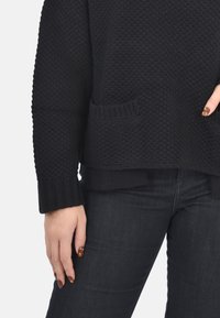 Maglione nero lavorato a maglia con un motivo testurizzato, ampie maniche a costine e una tasca frontale. Abbinato a jeans scuri.