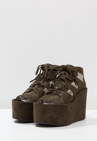 Bottes plateformes vert olive foncé avec une finition en daim, présentant un bout fermé, des côtés ouverts et un laçage. Talon en wedge épais.