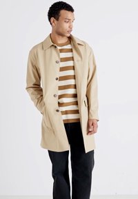 Beige színű trench coat, galléros nyakkal, elülső gombokkal és oldalsó zsebekkel, egy barna-fehér csíkos póló fölött viselve, fekete nadrággal párosítva.