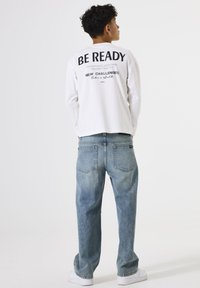 Witte lange mouw t-shirt met zwarte tekst op de achterkant, gecombineerd met losse lichtblauwe jeans. Witte sneakers maken de outfit compleet.