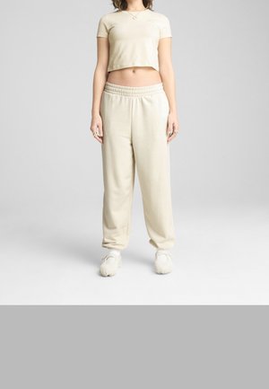 Person iført beige cropped kortærmet top, matchende højtaljede sweatpants og lyse teksturerede slip-on sko, stående med arme langs siderne.