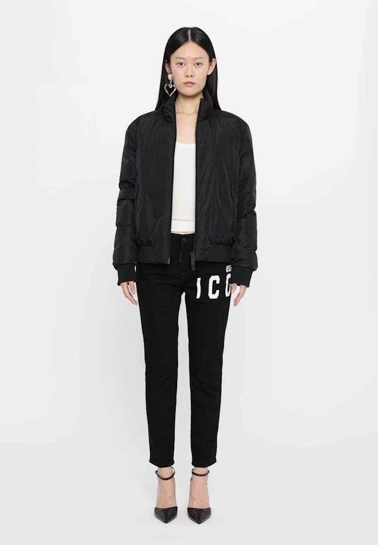 Veste bomber noire à la texture lisse, fermeture éclair, et poignets côtelés, associée à un pantalon noir avec des lettres blanches. Chaussures à talons noires.