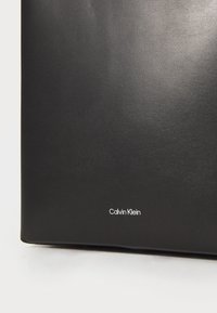 Czarna skórzana torebka o gładkiej fakturze i minimalistycznym designie, z białym logo "Calvin Klein" umieszczonym w dolnym prawym rogu.