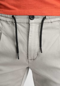 Lichtgrijze broek met een elastische tailleband, zwarte trekkoord en een knoopsluiting. Kenmerkt zich door een gladde stof en zijzakken.