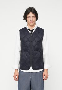 Μπλε καπιτονέ γιλέκο με φερμουάρ, που διαθέτει διαμαντένιο μοτίβο και απλό σχέδιο. Φορεμένο πάνω από ένα λευκό πουκάμισο και με μια μαύρη γραβάτα.