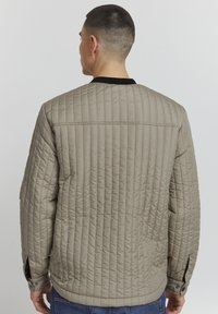 Giacca trapuntata beige con vestibilità rilassata, dotata di colletto a costine nero e fessure laterali ai polsini per un comfort aggiuntivo.