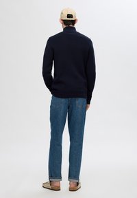 Maglione in maglia blu navy, jeans in denim azzurro e sandali chiari. Il modello indossa un cappello beige, girato di spalle rispetto alla macchina fotografica.