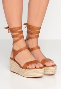 Bruna lädersandaletter med kilklack och ankelband, med en texturerad juteplattform och öppen tådesign. Remmarna är släta och platta.