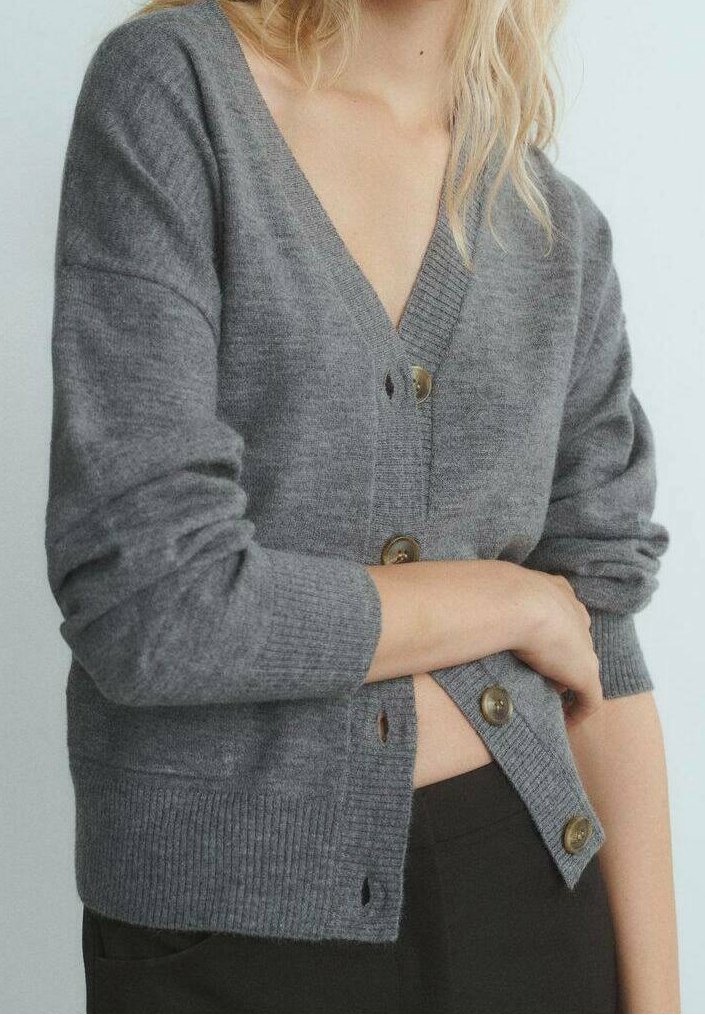 Femme portant un cardigan gris boutonné avec les manches retroussées, une main ajuste les boutons du bas, sur un fond clair uni.