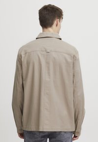 Beige cotton shirt jacket with a back yoke, buttoned cuffs, and a small logo label near the collar. Features a relaxed fit and straight hem.