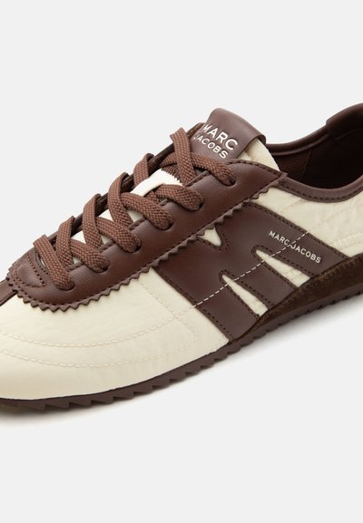 Αθλητικό παπούτσι χαμηλού προφίλ Marc Jacobs σε ανοιχτό κρέμ και καφέ δέρμα με καφέ κορδόνια που ταιριάζουν και εμπορικά σήματα στη γλώσσα και το πλάι.