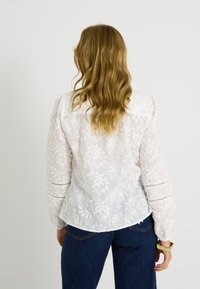 Blusa blanca bordada con mangas largas, patrón floral, textura transparente y detalles de encaje en las mangas, combinada con jeans azules.