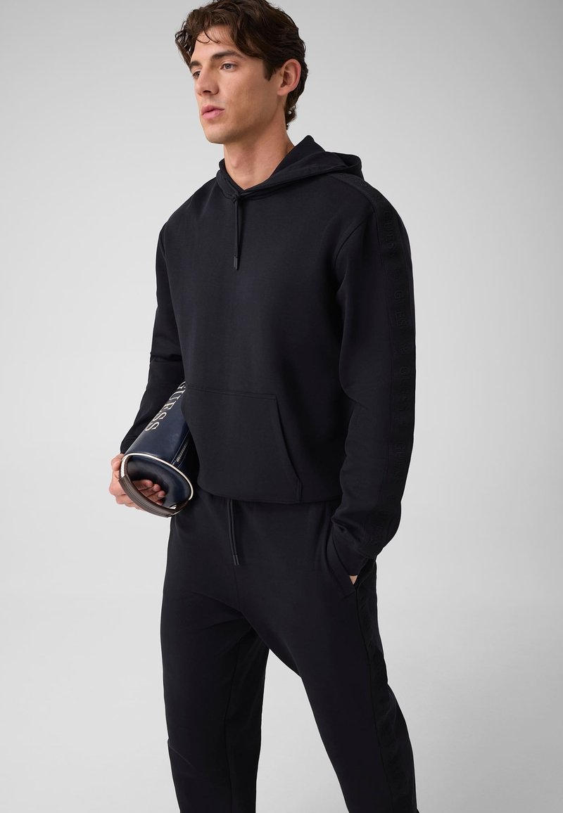 Homme en sweat à capuche noir et pantalon de jogging tenant un magazine roulé, debout contre un fond uni gris clair.