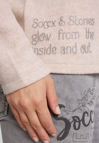 Maglione rosa chiaro, dalla texture morbida, con testo in glitter argento che leggono "Soccx & Stories brillano dall'interno e dall'esterno" sul davanti.