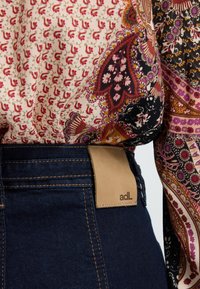 Wzorzysta bluzka z kwiatowymi i paisleyowymi motywami w kremowym, czarnym i czerwonym kolorze, zestawiona z ciemnoniebieskimi dżinsami z brązową skórzaną metką.