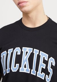 Černé bavlněné tričko s kulatým výstřihem, na kterém je v tučném modrém písmu obrysem v bílé barvě napsáno "DICKIES". Hladká textura, ležérní design.
