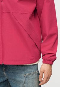 Gros plan d'une personne portant une veste zippée rose vif avec des poignets élastiques et un jean bleu délavé avec une poche avant visible.
