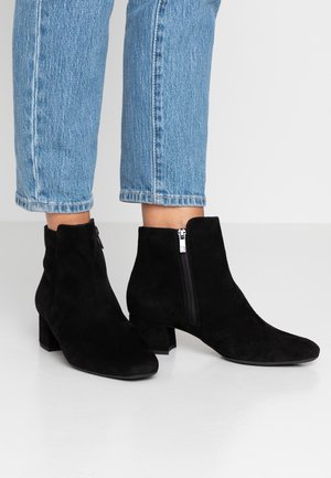 Pieds portant des bottines en daim noir à talons blocs avec fermetures éclair latérales, associées à un jean court bleu clair.