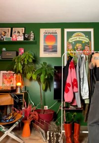 Pièce aux murs verts avec des affiches, une grande plante d’intérieur, un porte-manteau tenant des vestes, une paire de bottes rouges et une personne tatouée de dos.