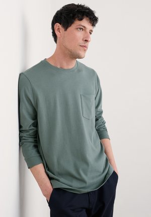 Olivenfarbener Langarm-T-Shirt aus weichem Stoff mit rundem Ausschnitt und einer Brusttasche. Kombiniert mit dunklen Hosen.