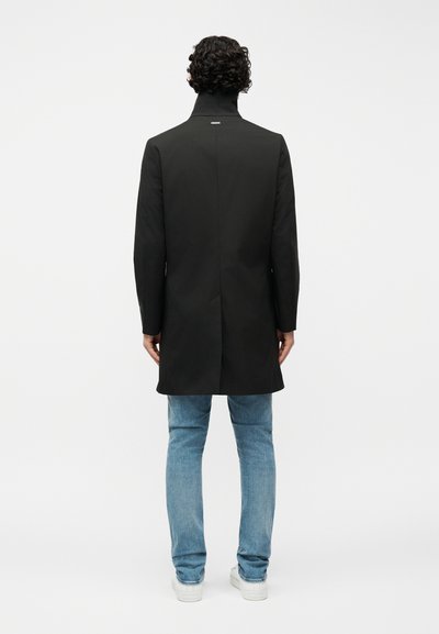 Άνδρας με σγουρά μαλλιά, φορώντας ένα μαύρο παλτό μεσαίου μήκους, μπλε τζιν και λευκά αθλητικά παπούτσια, στέκεται με την πλάτη προς τα εμπρός σε απλό φόντο.