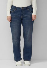 Jean en denim bleu foncé avec une coupe droite, cinq poches et des coutures visibles. Porté avec des baskets blanches.