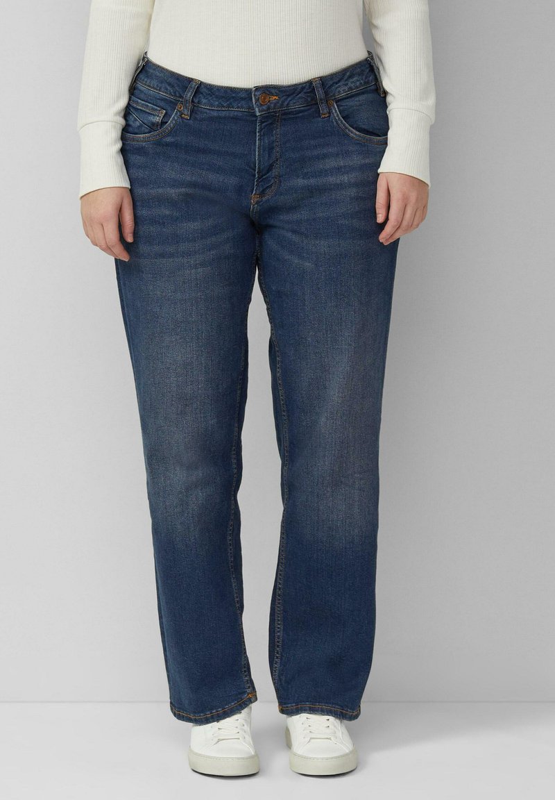 Jean en denim bleu foncé avec une coupe droite, cinq poches et des coutures visibles. Porté avec des baskets blanches.