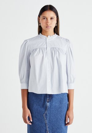Blouse rayée bleu clair en coton, avec un col haut à volants, des manches bouffantes et un devant à boutons. Associée à une jupe en denim.