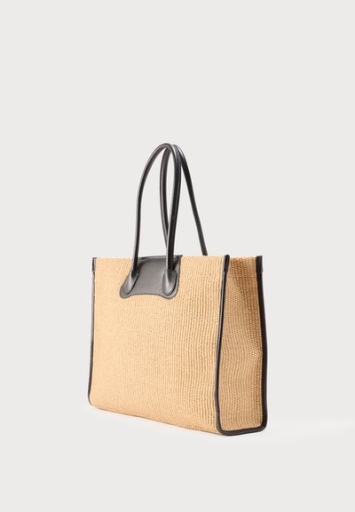 Μεγάλη μπεζ υφαντή τσάντα tote με μαύρες δερμάτινες λαβές, φινιρίσματα και γωνιακές λεπτομέρειες, εμφανίζεται σε απλό ανοιχτό γκρι φόντο.