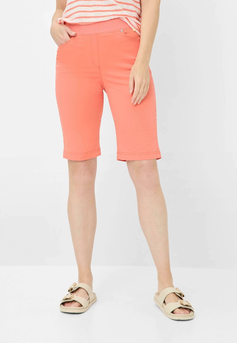 Shorts corail mi-cuisse avec une texture lisse, dotés d'un ourlet cousu et de deux poches frontales. Associés à des sandales à glissière beiges avec des nœuds décoratifs.