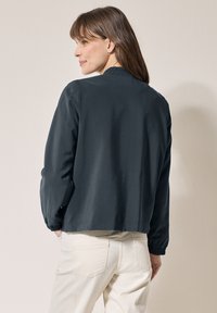 Mörkgrå lättviktsjacka med elastiska manschetter och rynkad halsringning, bärs över ljusbeiga byxor. Slät textur och avslappnad passform.